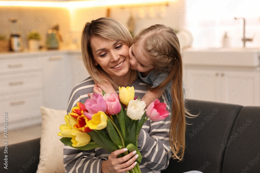 Little daughter kissing and congratulating her mom with Mother`s Day at home. Woman holding bouquet of beautiful tulips