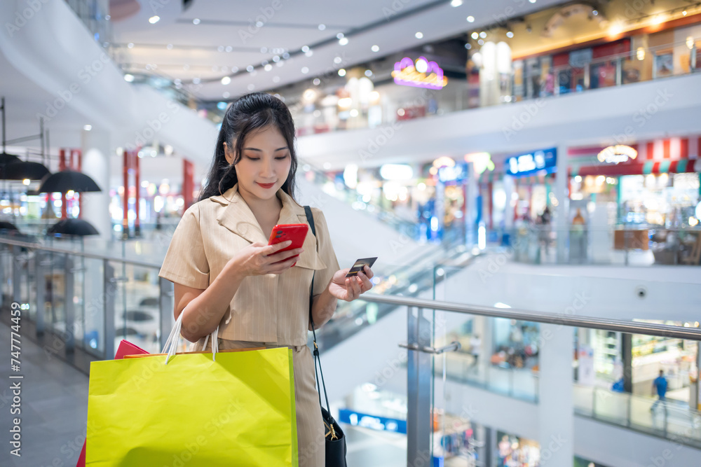 young woman in smart casual wear carrying many paper shopping bags shopping online on mobile phone using credit card at shopping center young woman in smart casual wear carrying many paper shopping bags shopping online on mobile phone using credit card at shopping center
