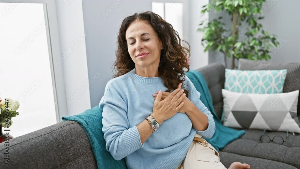 Grateful middle age hispanic woman with heart-filled gesture, sitting at home on the sofa, beaming health and happiness Grateful middle age hispanic woman with heart-filled gesture, sitting at home on the sofa, beaming health and happiness