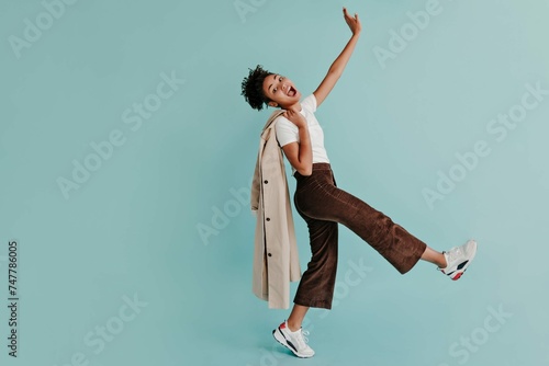 Refined African Girl Standing One Leg Waving Hand Turquoise Background Full Length View Smiling Black Woman With Trench Coat photo
