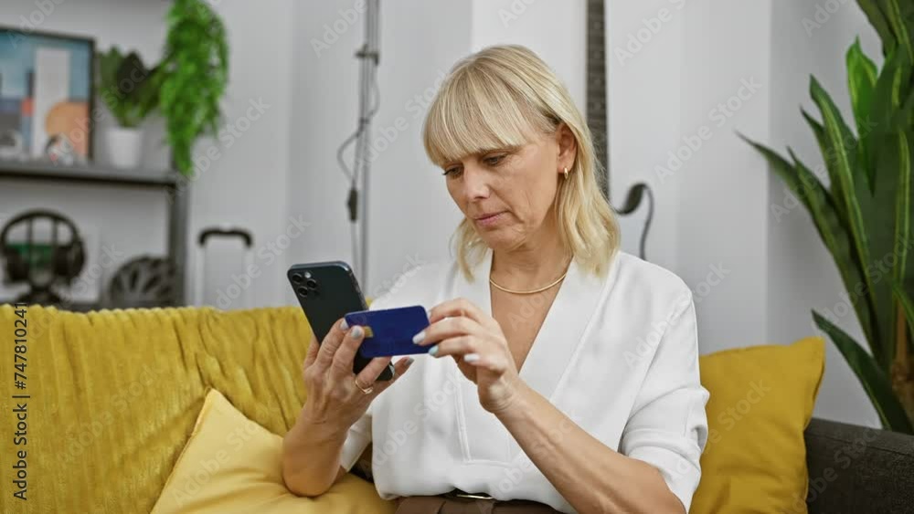 A blonde woman examines a credit card while using a smartphone in a modern living room. A blonde woman examines a credit card while using a smartphone in a modern living room.