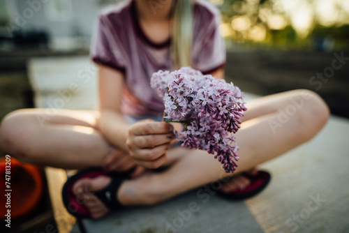 Φωτογραφία Girl sitting cross legged holding out lilacs on summer evening.