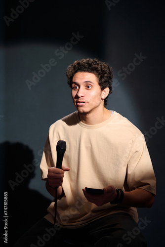 Young comedian, actor or showman with microphone and smartphone sitting on stage in front of audience during monologue