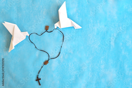 Two white dove origami carrying rosary or scapular in sky blue background. 