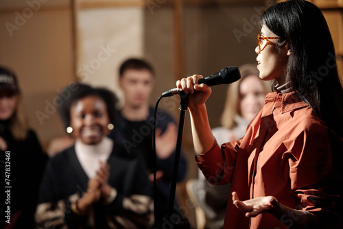 Young comedian of stand up show standing against audience and pronouncing monologue in microphone during performance