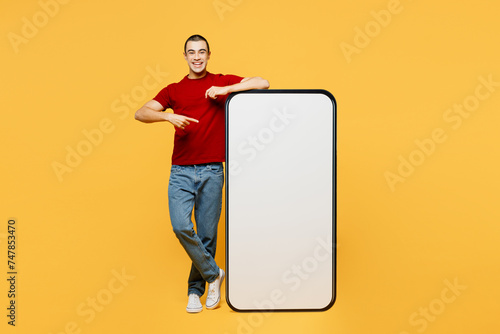 Full body fun young middle eastern man he wear red t-shirt casual clothes point index finger on big huge blank screen mobile cell phone smartphone with area isolated on plain yellow orange background.