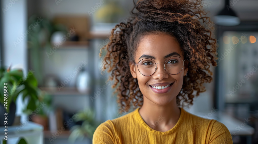 A cheerful young woman with curly hair wearing a yellow sweater and glasses in a cozy interior