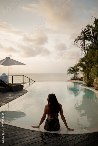 Rear view slim brunette woman in black swimsuit sitting on the edge of infinity swimming pool and enjoy sunset by the sea. Tropical vacation concept