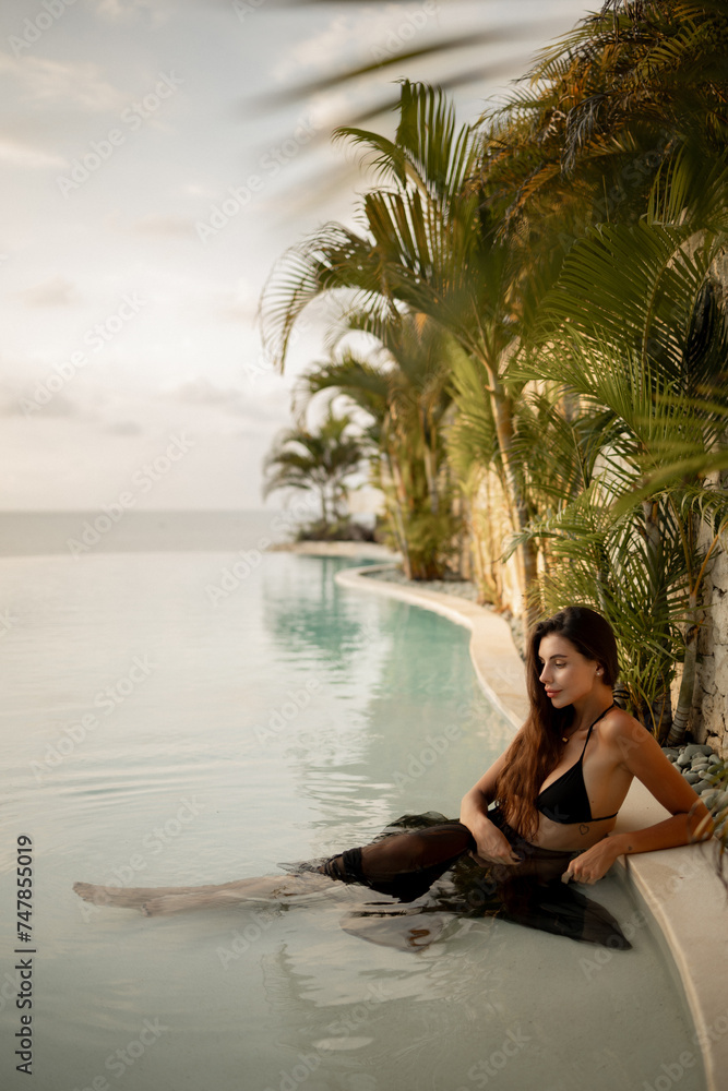 Rear view slim brunette woman in black swimsuit sitting  in water in infinity swimming pool and enjoy sunset by the sea. Tropical vacation concept