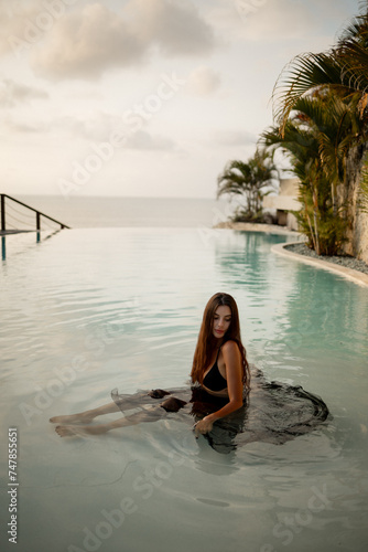 Young pretty attractive stylish slim sporty caucasian woman girl sitting in swimming pool wearing black transparent maxi skirt swimwear . Luxury resort vacation travel day concept