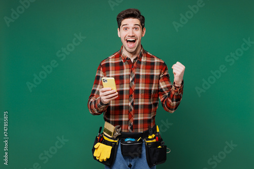 Young winner employee laborer handyman man wearing red shirt hold use mobile cell phone isolated on plain green background. Instruments accessories for renovation apartment room. Repair home concept. photo