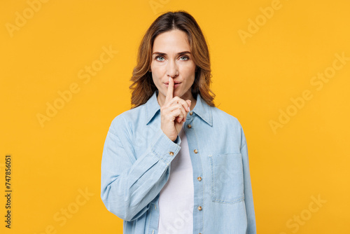 Young woman she wearing blue shirt white t-shirt casual clothes say hush be quiet with finger on lips shhh gesture look camera isolated on plain yellow background studio portrait. Lifestyle concept.