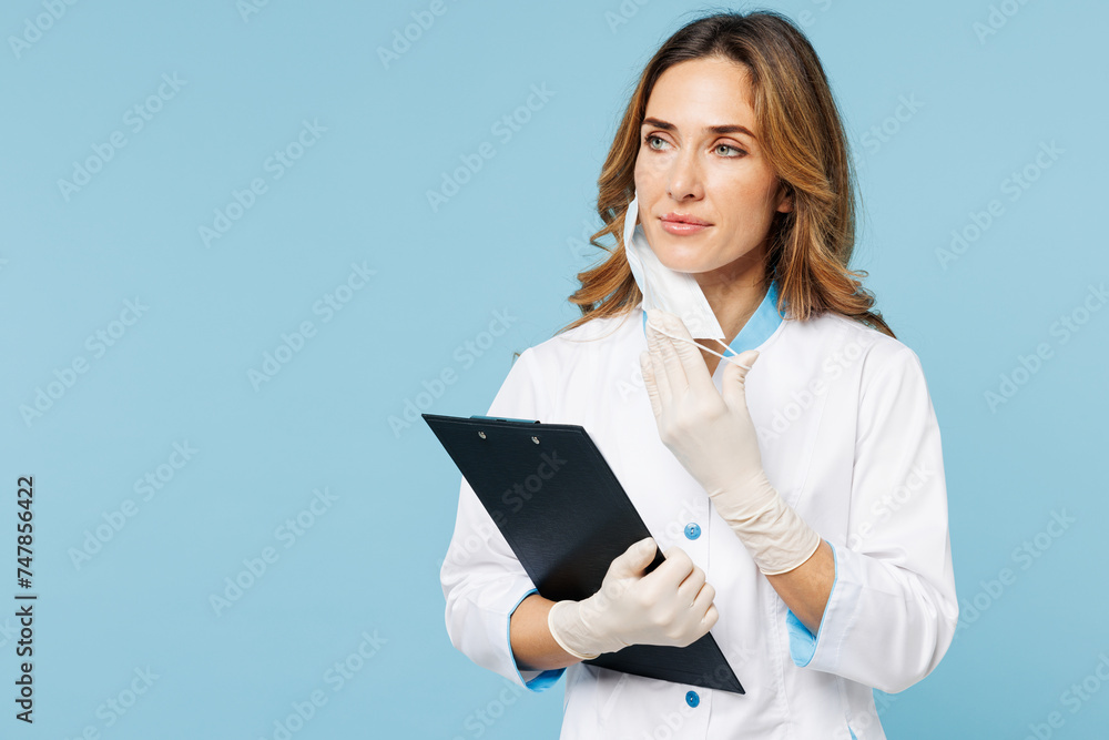 Female doctor woman wears white medical gown suit work in hospital clinic office hold clipboard with documents look aside take off mask isolated on plain blue background. Health care medicine concept. Female doctor woman wears white medical gown suit work in hospital clinic office hold clipboard with documents look aside take off mask isolated on plain blue background. Health care medicine concept.