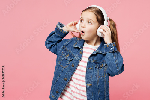 Fototapeta Naklejka Na Ścianę i Meble -  Little child shocked cute kid girl 7-8 years old wear denim shirt listen to music in headphones look aside on area isolated on plain pastel pink background. Mother's Day love family lifestyle concept.