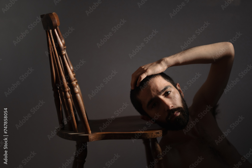 A shirtless, bearded, multiracial Asian, Jewish, and White man leans his head against a wooden chair, and turns to look at the camera with his hand on his head. He looks serious and vulnerable.