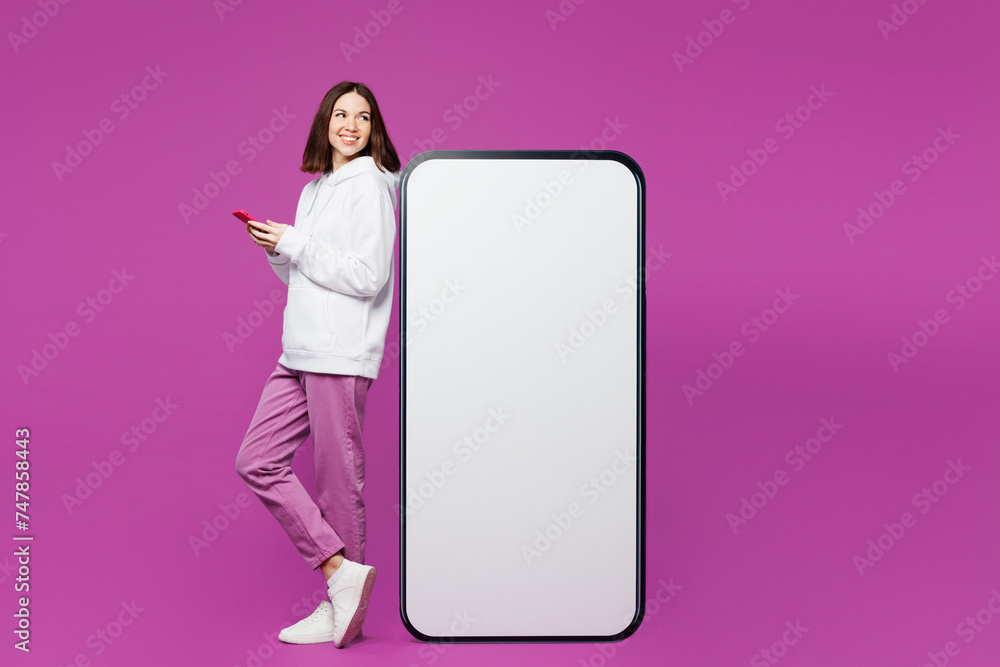 Full body side view young woman wear light hoody casual clothes stand near big huge blank screen mobile cell phone with area use smartphone isolated on plain purple color background Lifestyle concept Full body side view young woman wear light hoody casual clothes stand near big huge blank screen mobile cell phone with area use smartphone isolated on plain purple color background Lifestyle concept