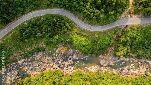 Aerial view of a winding road through lush green tropical forest, with a river and rocky terrain visible, suitable for adventure travel and environmental themes