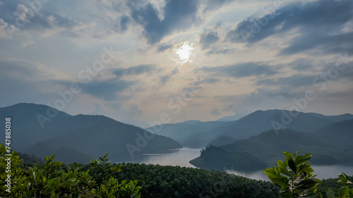 Scenic view of a tranquil lake nestled among rolling hills under a hazy sky, with sun rays peeking through clouds, ideal for nature-themed backgrounds with space for text, Earth day photo