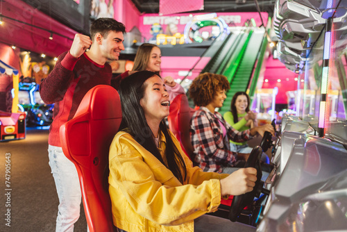 Asian woman playing with her multiethnic friends in racing games in an arcade game center while her friends celebrate and cheer