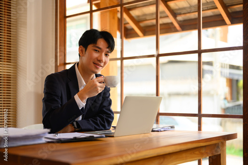 Young businessman in black suit sitting at desk with laptop and drinking coffee