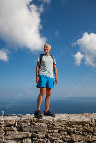 Caucasian senior male standing on the stone wall with blue sea and cloudy sky in a background (Amorgos, Cyclades/Greece)