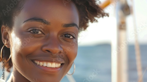 A smiling woman with curly hair wearing a hoop earring and a septum piercing set against a blurred background of a boat and water.