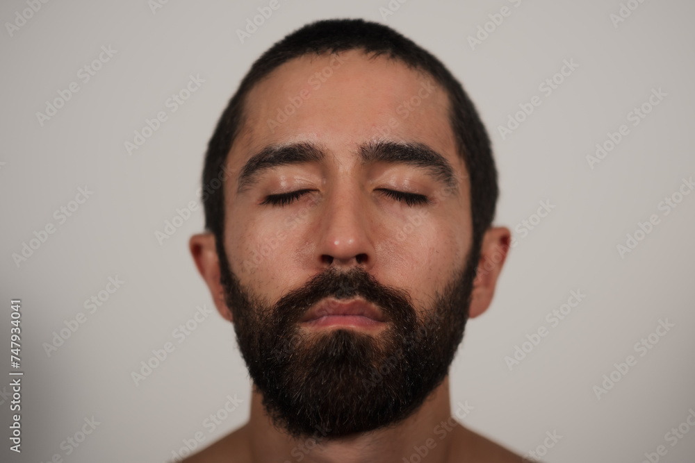 A closeup studio portrait of a man with closed eyes, posing shirtless against a white background. Soft lighting draws attention to his dark beard, strong eyebrows, and the hairline of his trim hair. A closeup studio portrait of a man with closed eyes, posing shirtless against a white background. Soft lighting draws attention to his dark beard, strong eyebrows, and the hairline of his trim hair.