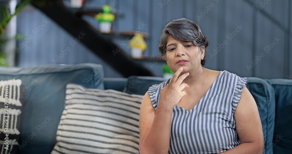 Asian old elder sad stress tired tense female patient sitting alone on sofa couch feel bad at indoor room home. Indian lonely aged gen x adult older upset woman lady think family suffer health issue