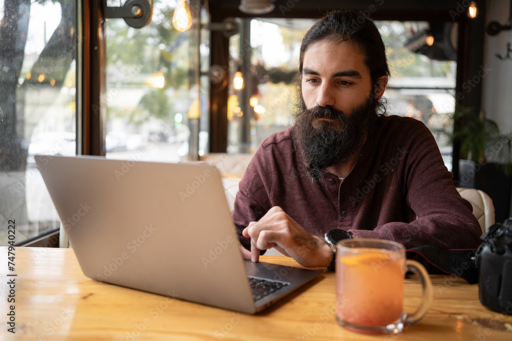 Pensive handsome male blogger with long beard working uses social networks watch multimedia files sitting in cafe with equipment and laptop computer Pensive handsome male blogger with long beard working uses social networks watch multimedia files sitting in cafe with equipment and laptop computer