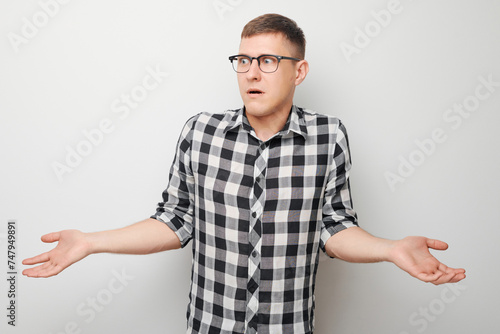 Confused young man in checkered shirt shrugging shoulders on white background expressing uncertainty