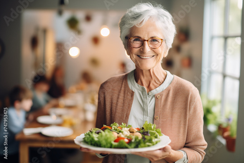Aged woman smiling happily and holding a healthy vegetable salad bowl on blurred kitchen background  with copy space.