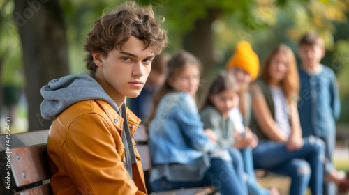 A young teenager sits contemplatively on a park bench, feeling isolated with blurred figures in the background during a sunny autumn day. bullying among teenagers photo