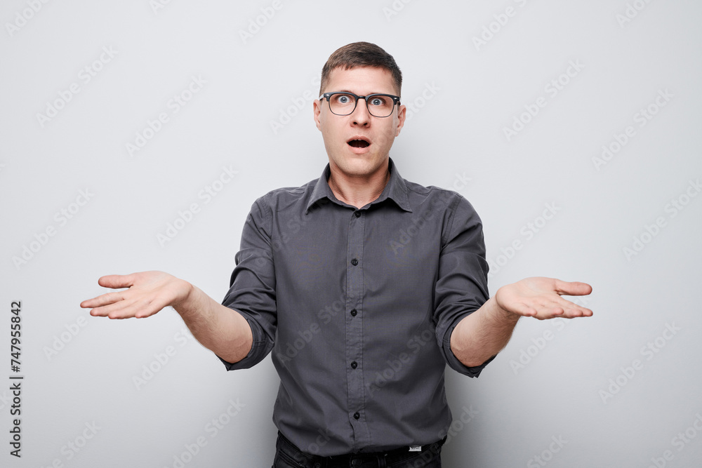 Confused young man in glasses and grey shirt, shrugging with hands outstretched on plain background Confused young man in glasses and grey shirt, shrugging with hands outstretched on plain background