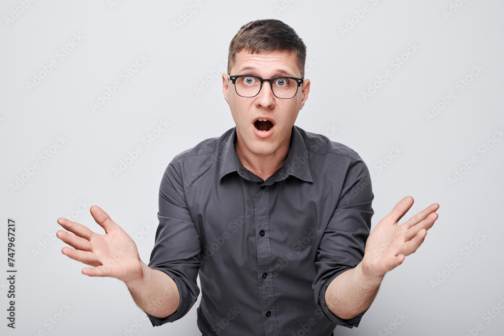 Fototapeta premium Surprised young man with glasses hands raised and mouth open in shock against a white background.