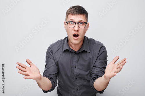 Surprised young man with glasses hands raised and mouth open in shock against a white background.