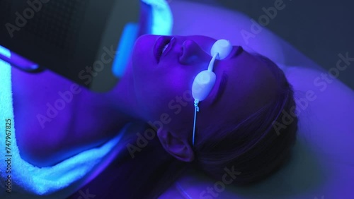 Closeup tracking shot of female client having blue LED light facial photodynamic therapy treatment in beauty salon. Young woman lying during non-invasive type of phototherapy. Shooting in slow motion. photo