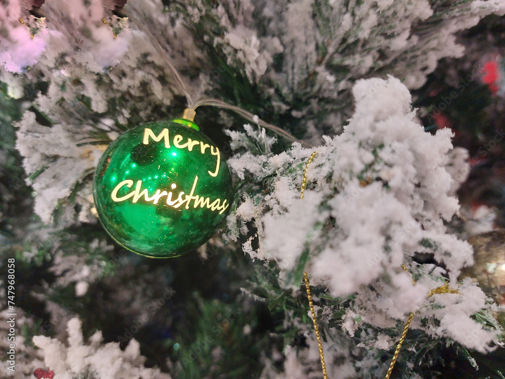 Merry Christmas and Happy New Year! A green Christmas ball with the message Merry Christmas hangs on a branch of a fake snow-covered Christmas tree.