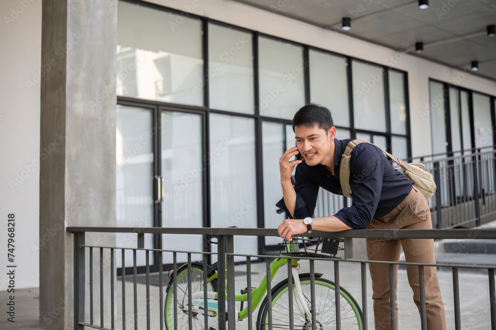 A young Asian businessman rides a bicycle to work. Standing outside the office talking on the phone and talking about business. Concept of reducing energy and reducing air pollution. A young Asian businessman rides a bicycle to work. Standing outside the office talking on the phone and talking about business. Concept of reducing energy and reducing air pollution.