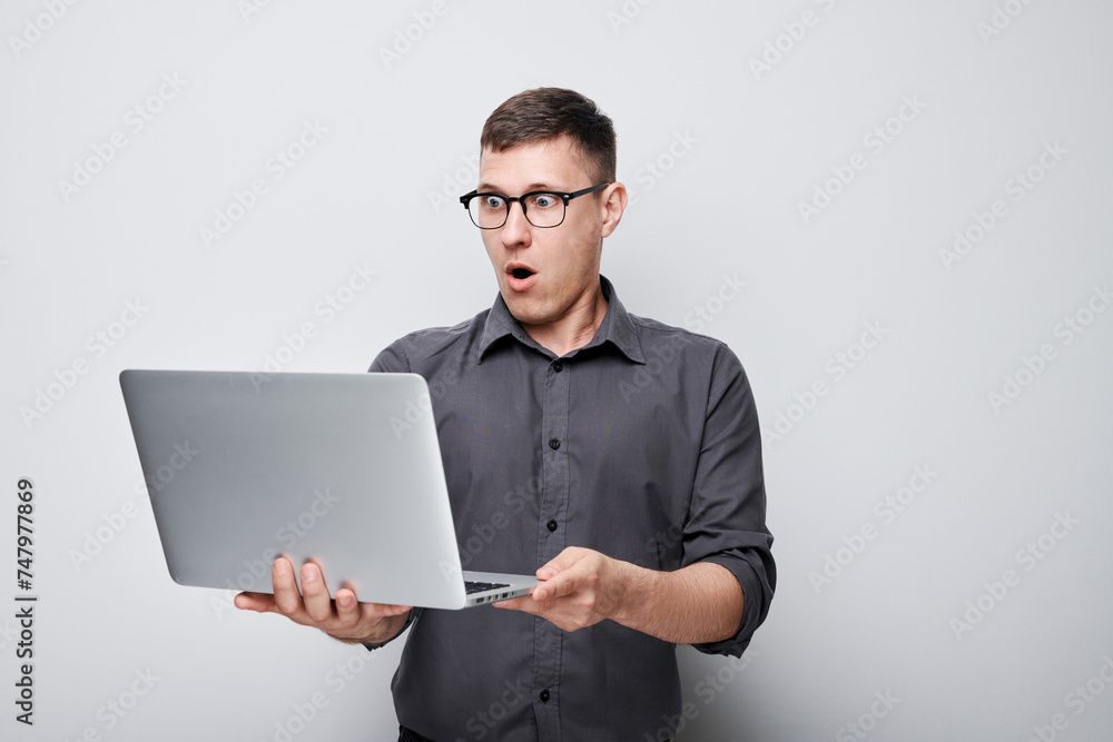 Surprised young man in glasses holding laptop with open mouth expression isolated on gray background Surprised young man in glasses holding laptop with open mouth expression isolated on gray background