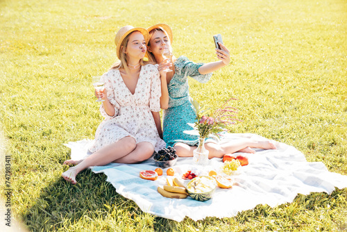Two young beautiful smiling hipster female in summer sundress and hats. Carefree women making picnic outside. Positive models sitting on plaid on grass, drinking champagne,  eating fruits, take selfie