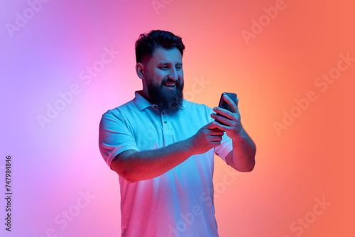 Portrait of young man holding smartphone and smiling lookin to screen in neon light against vibrant gradient studio background. Video call. Concept of human emotions, connection in distance. Ad photo