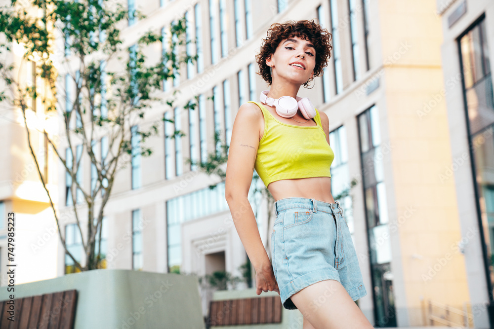 Young beautiful smiling hipster woman in trendy summer clothes. Carefree woman with curls hairstyle, posing in the street at sunny day. Positive model outdoors. Listens music at her headphones on neck