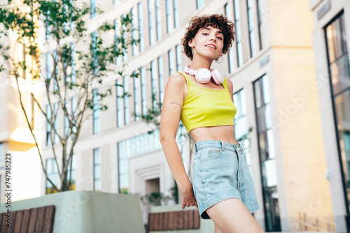 Young beautiful smiling hipster woman in trendy summer clothes. Carefree woman with curls hairstyle, posing in the street at sunny day. Positive model outdoors. Listens music at her headphones on neck