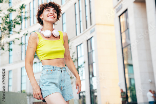 Young beautiful smiling hipster woman in trendy summer clothes. Carefree woman with curls hairstyle, posing in the street at sunny day. Positive model outdoors. Listens music at her headphones on neck