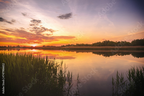 Sunset or sunrise above the pond or lake at spring or early summer evening or morning with cloudy sky and reed grass. Springtime landscape. Water reflection. Vintage film aesthetic.