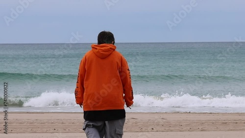 Mujer joven con sudadera naranja y estilo alternativo caminando hacia la orilla en la Playa de San Juan de Alicante, España photo