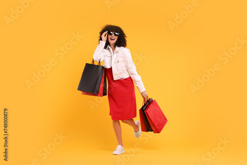 Happy young woman with shopping bags and stylish sunglasses on yellow background
