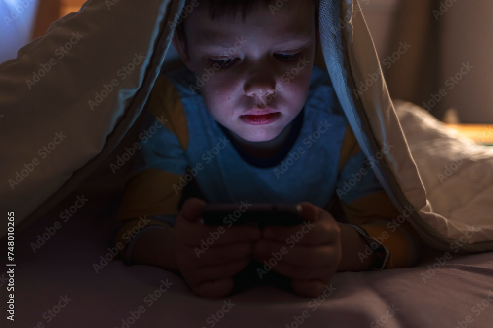 Boy playing games on mobile phone while lying on bed in bedroom under ...