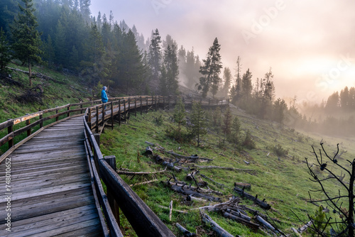 Man standing on a wooden boardwalk looking down a valley filled with fog from nearby hot springs brightly illuminated by the rising sun, Mud Volcano, Yellowstone National Park, Wyoming