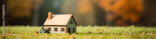 Banner, concept design for real estate agency, purchasing a plot of land and building a house. Miniature wooden model of a house standing in green grass on the background of an autumn forest.